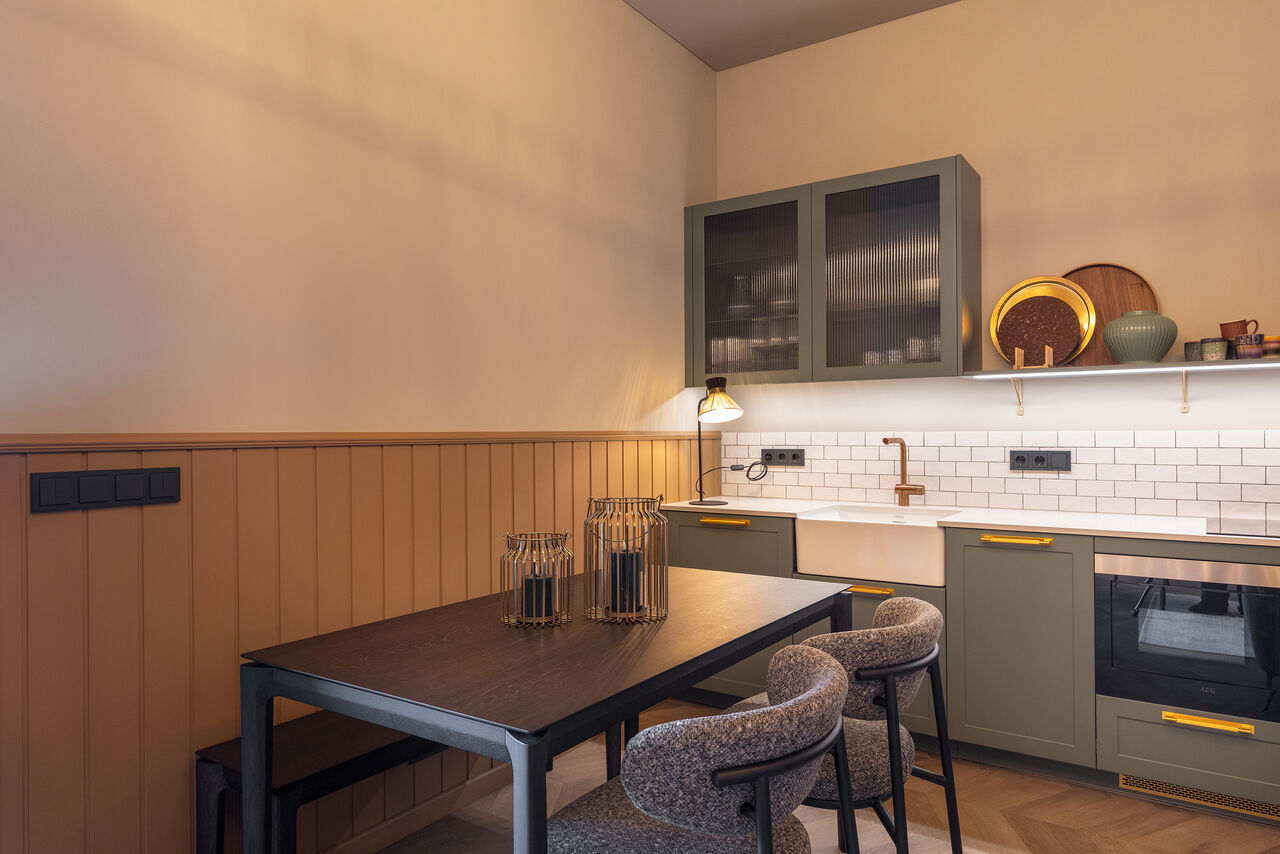A dining table in front of a sage-green kitchen with open shelves