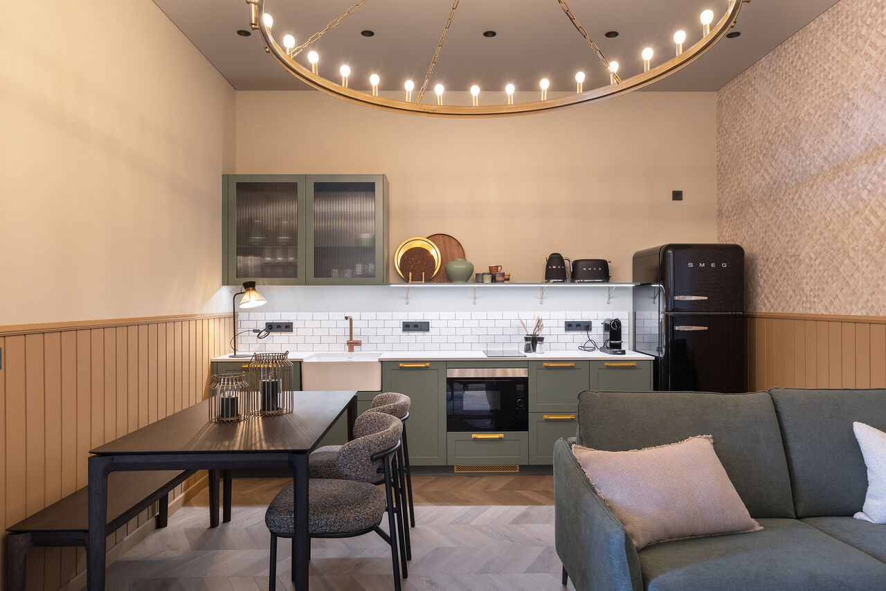 A one-bedroom living space with a dining table, sage sofa and kitchen under a brass ring chandelier